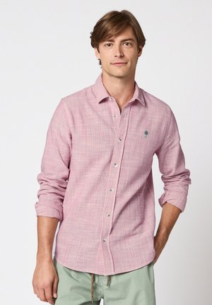 Joven vestido con camisa de botones a rayas rosas con las mangas remangadas y pantalones verde claro, de pie frente a un fondo liso.