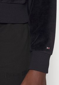 Felpa in velluto nero con polsini a coste, dotata di un piccolo logo ricamato sulla manica e testo in rilievo che recita "Tommy Hilfiger" sulla vita.