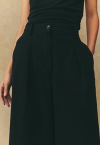 Pantaloni neri a gamba larga con vita alta, chiusura con bottone e tasche laterali. Realizzati in un tessuto leggero e morbido con dettagli plissettati.