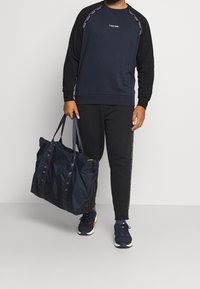 Sudadera azul marino con mangas raglán negras, logo en el pecho. Pantalones deportivos negros, llevando un bolso grande de mano azul oscuro, zapatillas azul marino.