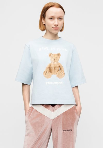 Νεαρό άτομο που φοράει ανοιχτό μπλε μπλουζάκι με εκτύπωση αρκουδάκι και το κείμενο «Bear in Mind», συνδυασμένο με ροζ παντελόνι βελούδου με λευκή λεπτομέρεια ζιγκ-ζαγκ.