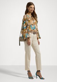 Blomsterbluse i beige, blå og brun med vide ærmer; parret med cremede jeans og turkise højhælede sandaler. Lille grå håndtaske.