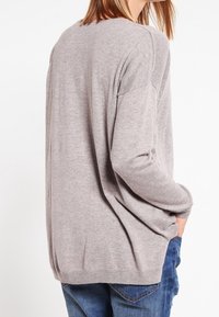 Femme portant un pull ample gris clair et un jean bleu, avec la main droite dans la poche, vue de dos sur un fond blanc.
