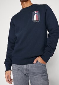 Granatowa bluza z okrągłym dekoltem, długimi rękawami i logo w kolorach czerwonym, białym i niebieskim na piersi. Miękka w dotyku.