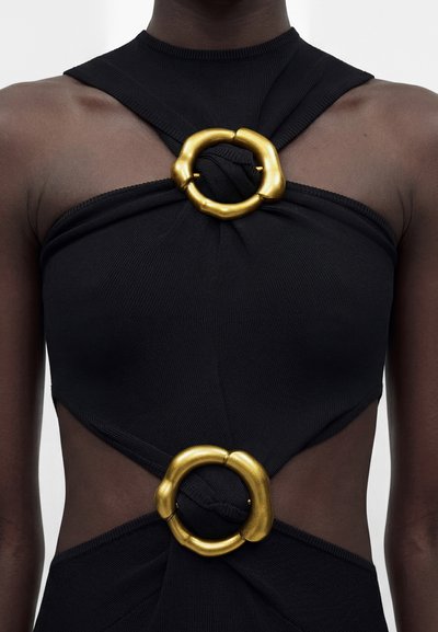 Μαύρο αμάνικο τοπ με δύο εμφανείς στρογγυλές χρυσές αγκράφες που σφίγγουν το ύφασμα στο στήθος και τη μέση σε άτομο με σκουρόχρωμο δέρμα.