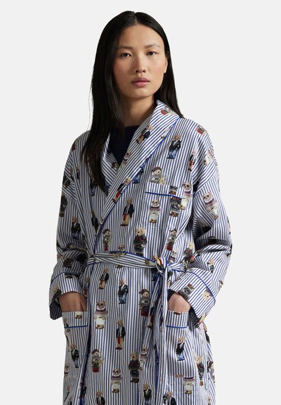 Jonge vrouw die een blauw-wit gestreepte kamerjas draagt met teddyberen in verschillende outfits, staand met handen in de zakken.