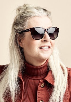 Bruine zonnebril met een glanzende afwerking, dikke monturen en donkere lenzen; gedragen met een geribbelde, hoge hals trui en een button-up jas.