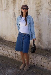 Camicia in denim blu chiaro con bottoni sopra una t-shirt bianca attillata, abbinata a pantaloni Bermuda in denim e scarpe beige senza lacci. Borsa nera.