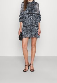 Černobílé šaty se vzorem zebry, dlouhými rukávy, našitou plisovanou horní částí a vícevrstvým volánovým lemem, doplněné černými sandály na podpatku a kabelkou.