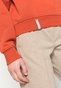 Oranges Sweatshirt mit gerippten Bündchen, kombiniert mit strukturierten beigefarbenen Hosen. Ein Logobutton ist am Saum des Sweatshirts sichtbar.