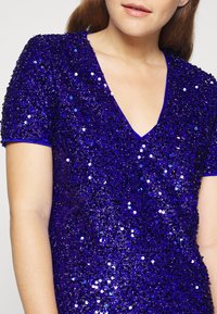 Lila klänning med V-hals, korta ärmar och en åtsittande design. Täckt av glänsande paljetter, med blå kant runt halsringningen och ärmarna.