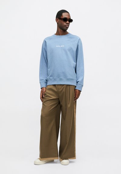 Sudadera azul con texto blanco, pantalones beige de pierna ancha y zapatos blancos. Modelo usando gafas de sol oscuras. Fondo blanco liso.