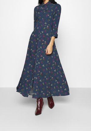 Kvinde iført en langærmet, marineblå blomstret maxikjole med bordeauxrøde ankelstøvler, stående mod en ensfarvet lys baggrund.
