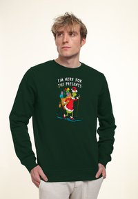 Giovane uomo che indossa una felpa verde scuro con il Grinch dello sci che porta regali e la scritta "Sono qui per i regali" sul davanti.