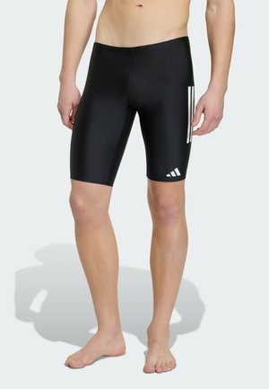 Mężczyzna w czarnych kąpielówkach do kolan z białymi bocznymi paskami i logo Adidas, stojący boso na jasnym tle.