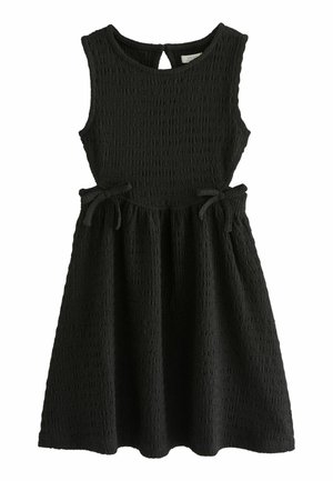 Enfant portant une robe noire sans manches texturée avec des liens latéraux et des sandales noires à boucle, debout devant un fond uni.