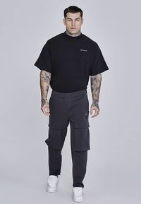 Zwarte oversized T-shirt gecombineerd met donkergrijze cargobroek met zijzakken, een klein logo en witte sneakers tegen een grijze achtergrond.
