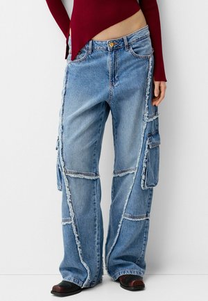 Osoba nosząca niebieskie jeansy cargo z denimu patchworkowego z surowymi krawędziami, czarne buty i bordowy top z długim rękawem, stojąca na białym tle.