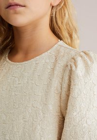 Bež obleka z teksturo, ki vključuje obliko napihnjenih rokavov. Tkanina ima cvetlični vzorec z diskretnim sijajem in okroglo ovratnico.