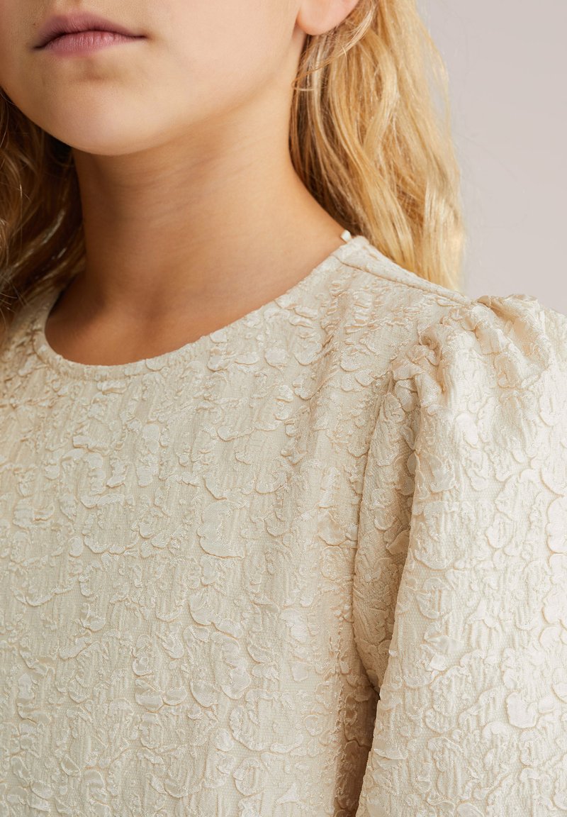 Bež obleka z teksturo, ki vključuje obliko napihnjenih rokavov. Tkanina ima cvetlični vzorec z diskretnim sijajem in okroglo ovratnico.