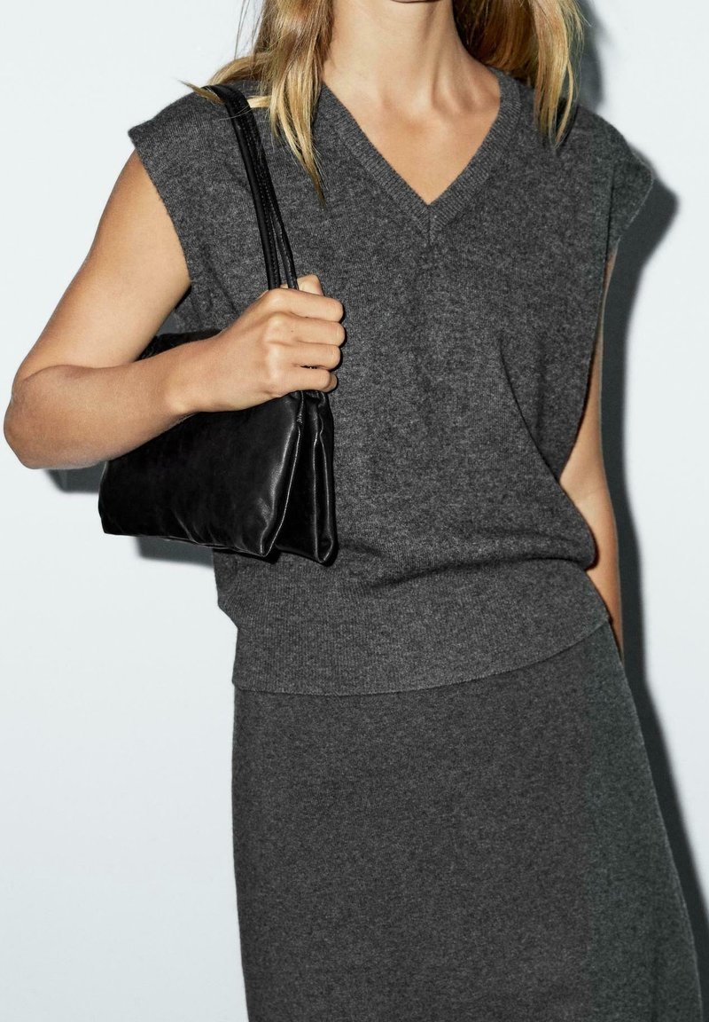 Femme portant un haut en maille sans manches gris foncé à col en V et une jupe assortie, tenant un petit sac à bandoulière en cuir noir sur son bras droit.