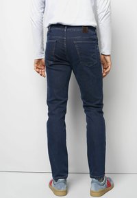 Jean en denim bleu foncé, coupe slim, avec cinq poches et des coutures contrastées. Portés avec des baskets colorées et une chemise blanche à manches longues.