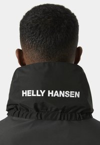 Černá, voděodolná bunda s vysokým límcem. Obsahuje bílé nápisy "HELLEY HANSEN" na zadní straně, hladký povrch a řasený detail límce.