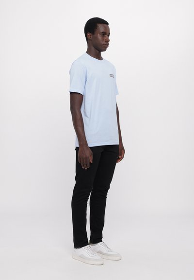 Lyseblå bomulds t-shirt med rund hals, sorte bukser og hvide sneakers. Enkelt design med et lille logo på brystet.