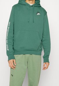 Grön hoodie i bomullsblandning, med en framficka, justerbar dragsko på huvan och vit tryckt logotyp på bröstet och ärmen.