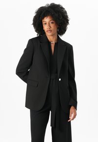 Zwarte op maat gemaakte blazer met een enkele zilveren knop, opgestikte kleppen en twee voorzakken; gecombineerd met een zwarte outfit voor een monochrome uitstraling.