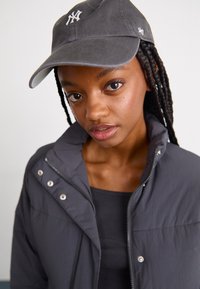 Doudoune grise à col montant et boutons-pression, portée sur un haut noir. Casquette de baseball grise avec un logo blanc.