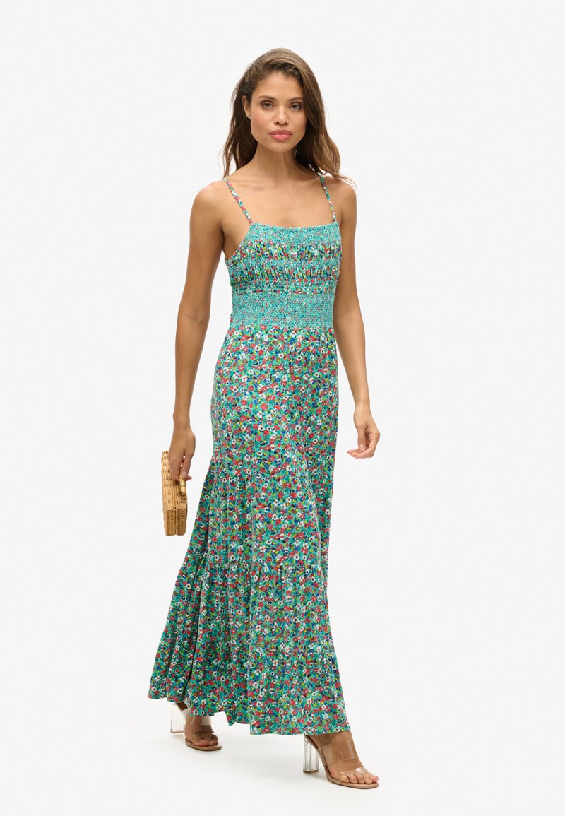 Kwiatowa sukienka maxi o turkusowym tle, z marszczonym topem, cienkimi ramiączkami i warstwową spódnicą. Modelka trzyma okrągłą wiklinową torbę.