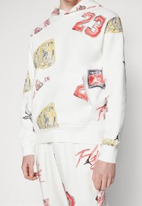 Sudadera blanca con capucha, adornada con gráficos multicolores y números. Cuenta con un bolsillo frontal y puños y dobladillo acanalados para mayor textura.