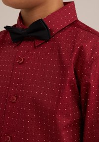 Röd skjorta med krage och vita prickar, med en matchande röd knapp och en svart fluga vid halsen. Slät textur, skräddarsydd passform.