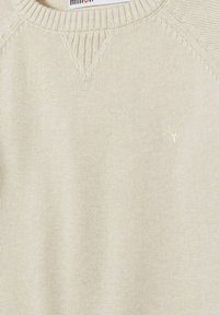 Ljusbeige stickad tröja med rund hals, ribbade muddarna och en liten broderad logotyp på bröstet. Slät textur, enkelt design.