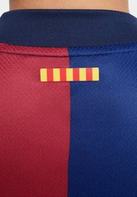 Odjeljak s dresom FC Barcelone s crvenim i plavim panelima; teksturirani materijal; žuta i crvena prugasta oznaka na ovratniku.