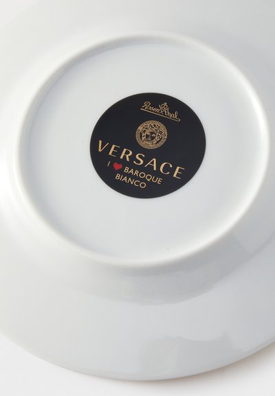 Bílý talíř s černou kruhovou nálepkou uprostřed, na které jsou zlaté a červené loga Versace Baroque Bianco a Rosenthal.