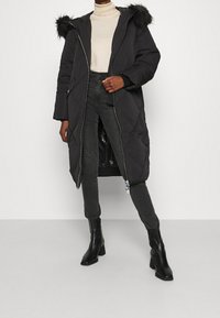 Černý prošívaný péřový kabát s kapucí lemovanou kožešinou, předním zipovým zapínáním a postranními kapsami. V kombinaci s tmavými džíny a černými botami na podpatku.