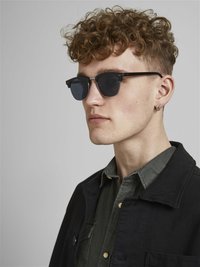 Sonnenbrille mit schwarzem Rahmen, dunklen Gläsern und silbernen Akzenten. Getragen von einer Person in einer schwarzen Jacke über einem dunkelgrünen Hemd. Lockige Haare.
