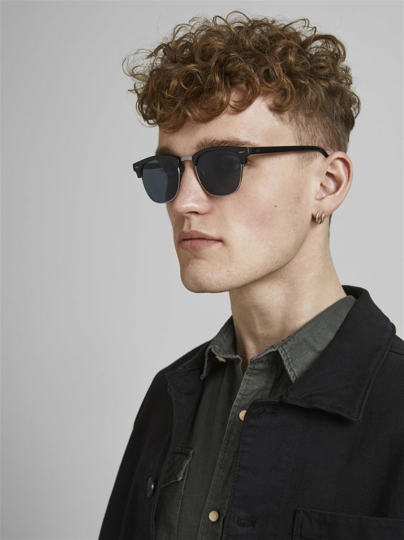 Sonnenbrille mit schwarzem Rahmen, dunklen Gläsern und silbernen Akzenten. Getragen von einer Person in einer schwarzen Jacke über einem dunkelgrünen Hemd. Lockige Haare.