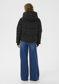 Schwarze Pufferjacke mit Kapuze, mit gesteppten Abschnitten und elastischen Bündchen, kombiniert mit weiten blauen Jeans. Glatte, matte Textur.