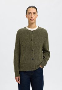 Olivgrön stickad cardigan med en fuzzig textur, rund halsringning och svarta knapplist, bärs över ett krämfärgat innerlager.