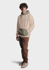 Sudadera color melocotón clara con un bolsillo verde, que presenta un logo en el pecho. Combinada con pantalones marrones y zapatos beige con detalles blancos.