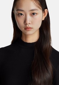 Detailní záběr na osobu s dlouhými tmavými vlasy a minimem make-upu, která má na sobě černý žebrovaný rolák. Přirozeně hladká pleť a neutrální výraz.