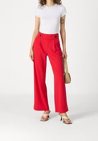 Czerwone szerokie spodnie z zakładkami z przodu, akcentami guzików i wysokim stanem, w połączeniu z białą dopasowaną koszulą i beżowymi sandałami na obcasie.