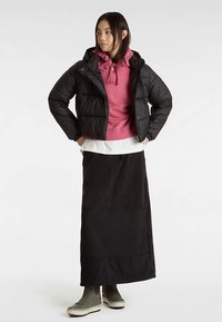 Veste noire matelassée sur un sweat à capuche rose, associée à une longue jupe noire et des bottes grises. La tenue présente une coupe décontractée et un design en couches.