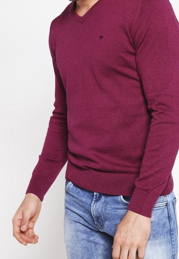 Pull-over à col en V couleur bordeaux, confectionné en tissu maille doux. Présente des poignets et une taille ajustés. Petit logo sur la poitrine. Porté avec un jean bleu.