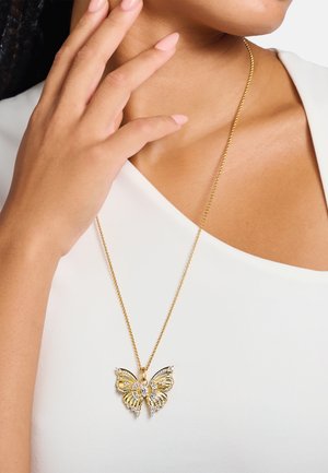 Goldener Schmetterlingsanhänger mit Strassakzenten an einer feinen Kette, getragen über einem weißen Oberteil, zeigt detaillierte Flügel.