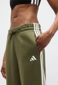 Spodnie dresowe w kolorze oliwkowozielonym z białymi paskami po bokach i logo Adidas. Posiadają elastyczny pas i boczną kieszeń. Gładka tekstura.