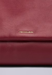 Burgundowa skórzana torebka o gładkiej fakturze. Posiada złote logo "TRUSSARDI" oraz czysty, minimalistyczny design.